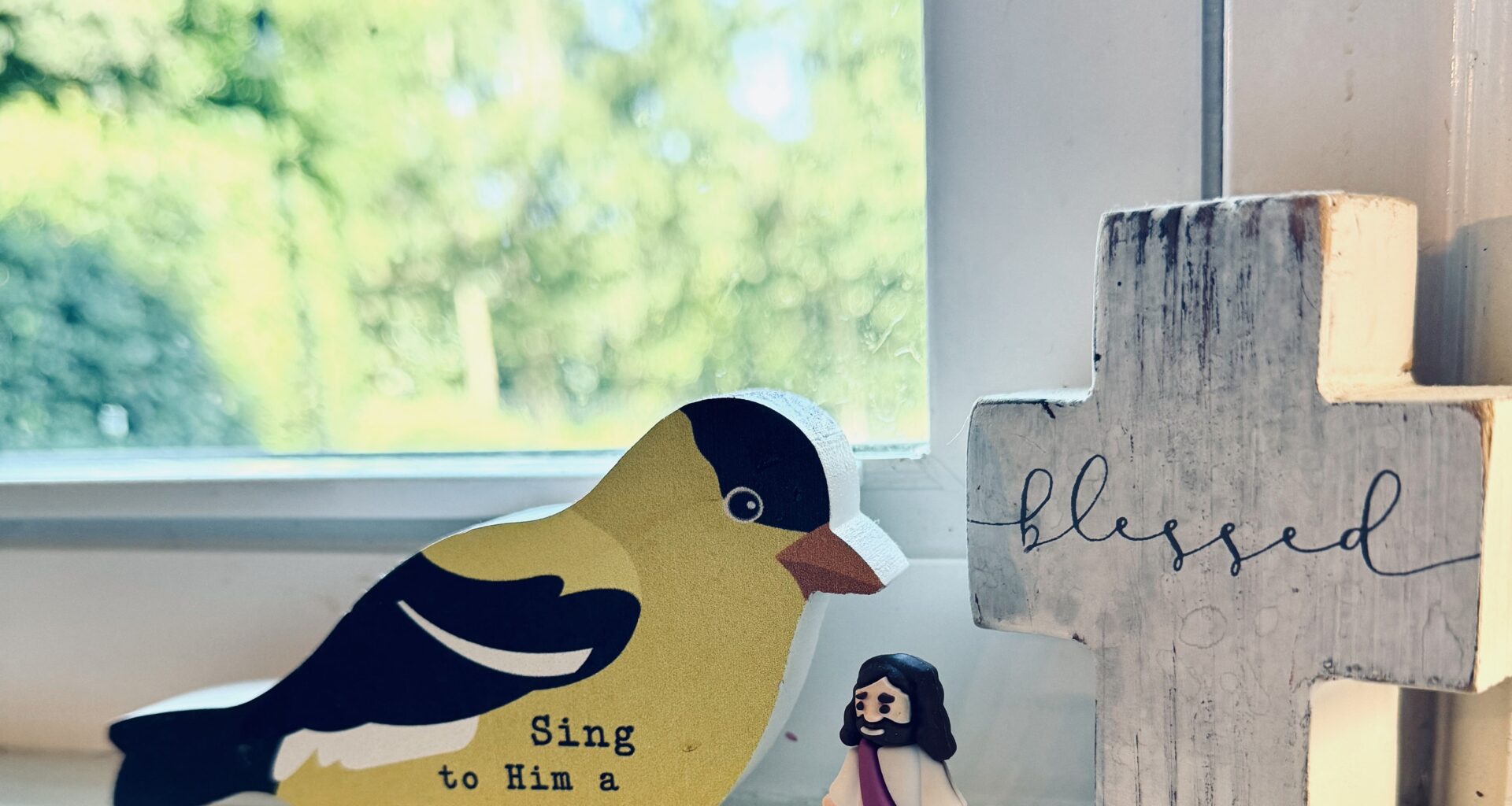 A trio of trinkets on Jamie's kitchen windowsill: a wooden yellow bird with black feathers with the words "Sing to him a new song," along with a miniature Jesus and a wooden cross with the word blessed in cursive printed across it.