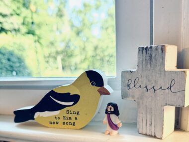 A trio of trinkets on Jamie's kitchen windowsill: a wooden yellow bird with black feathers with the words "Sing to him a new song," along with a miniature Jesus and a wooden cross with the word blessed in cursive printed across it.