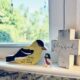 A trio of trinkets on Jamie's kitchen windowsill: a wooden yellow bird with black feathers with the words "Sing to him a new song," along with a miniature Jesus and a wooden cross with the word blessed in cursive printed across it.